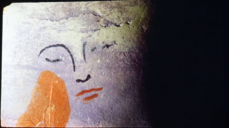 Still from moving image work showing a half-lit concrete wall with the outline of a colourful face painted on it.