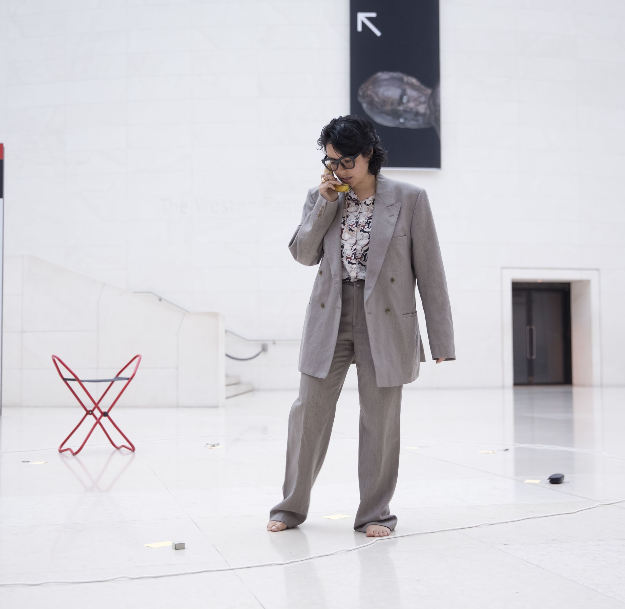 Photo by Rachel Cherry. Dre stands in a large grand foyer area pretending to be on the phone with a banana instead of a mobile. They wear an over-sized grey suit and thick rimmed glasses. A lone red folding stool is placed nearby in the background.