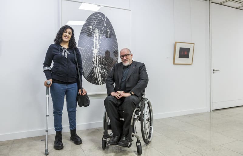 A white male wheelchair user and a young woman of South Asian heritage with a crutch are in a gallery space either side of a huge painting of a black oval on white canvas. They are looking at the camera and smiling