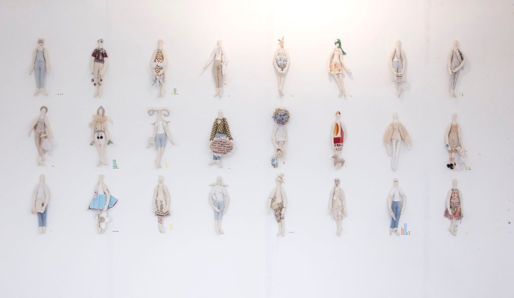 A white wall with three rows of eight fabric dolls pinned to it. The dolls are plush human figures with costumes and clothes on them.