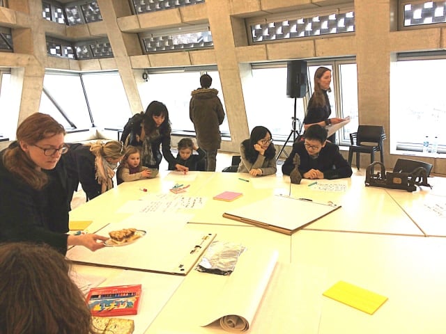 a group of people of varying ages and backgrounds sit around a large square table on which rest art objects for handling and inspection by participants