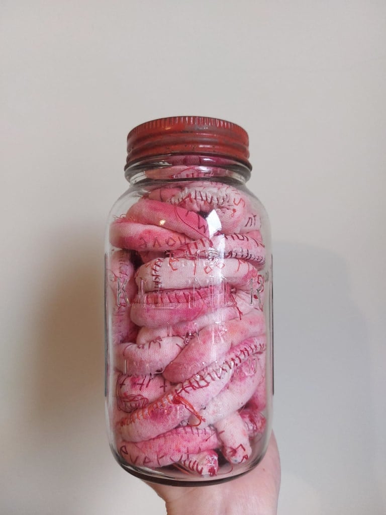 Infront of an off-white wall, a hand holds up a glass jar with a red lid. Inside is a long stuffed string of pink speckled and plump fabric with words embroidered into the fabric. Only parts of words are visible.