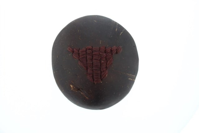 A circular brooch made from half a coconut shell, photographed from above. In the centre of the shell a triangle of dark down hair has been woven. The triangle is upside down which, as the artist describes, represents ‘the shape of the womb, to show 