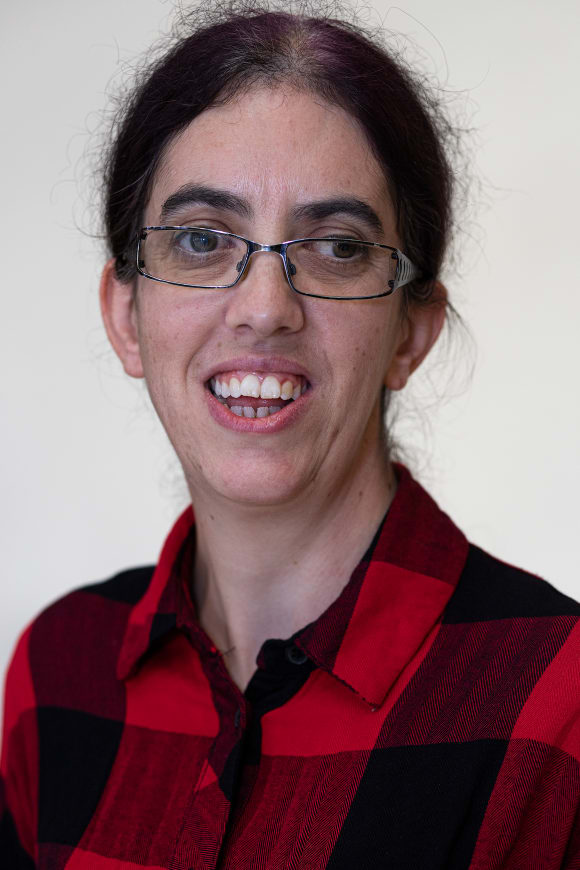 This colour portrait photograph of Emma, shows a white woman in her 40s with square rimmed glasses and dark wiry hair tied back behind her head. She looks slightly to the side of the camera and has a full smile with prominent teeth. Although the image is cropped and only her upper chest and head is visible, she gives the impression that she is tall and lean. She wears a red and black checked outfit with a collar, the top button undone. 