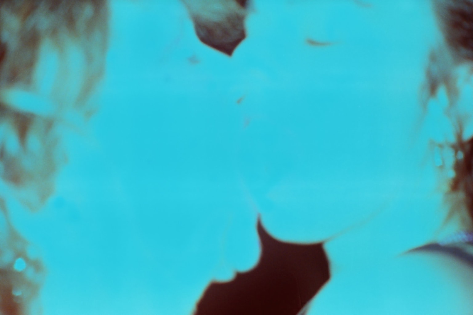 An over-exposed, cyan-tinted film photograph which shows a close up frame of two faces kissing.