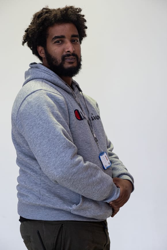 This colour photograph is a portrait of Abdul, a black man in his early 30s, who stands before a plain wall with his head turned to face the camera. His expression is soft, hinting at a slight smile. His grey hoodie and short afro hair is caught slig